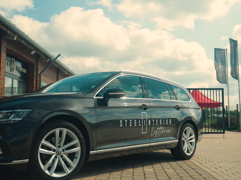 steel-interior-neuer-standort-2 Gray sedan with 'Steel Interior' branding parked outside a dealership under a partly cloudy sky.